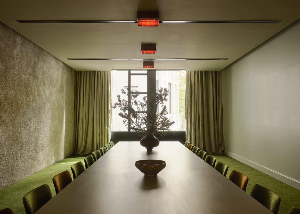 Minimalist dining room with a long table, green chairs, green carpeting, large window, and a vase with branches as centerpiece.