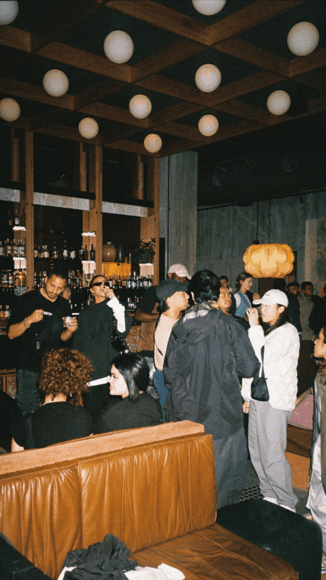 A group of people socializing and drinking in a dimly lit bar with shelves of bottles visible in the background.