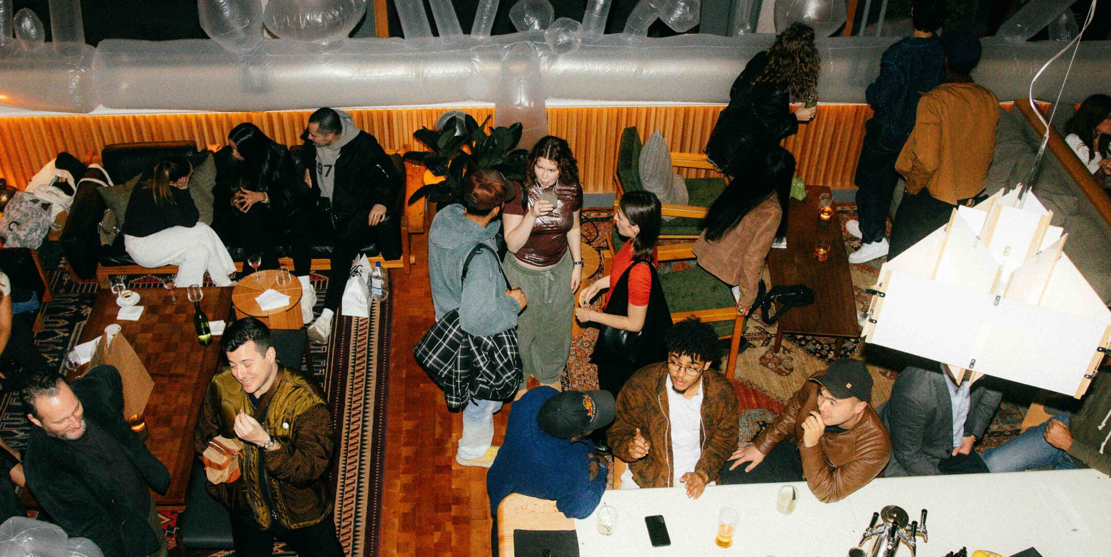 A group of people socializes in a lounge with drinks, couches, rugs, and warm lighting, viewed from above.